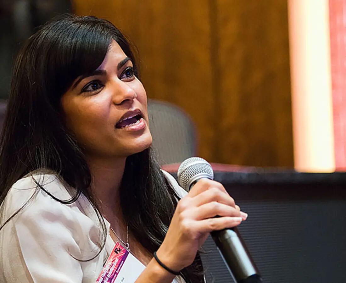 A woman with long black hair holding a microphone speaks at a past symposium